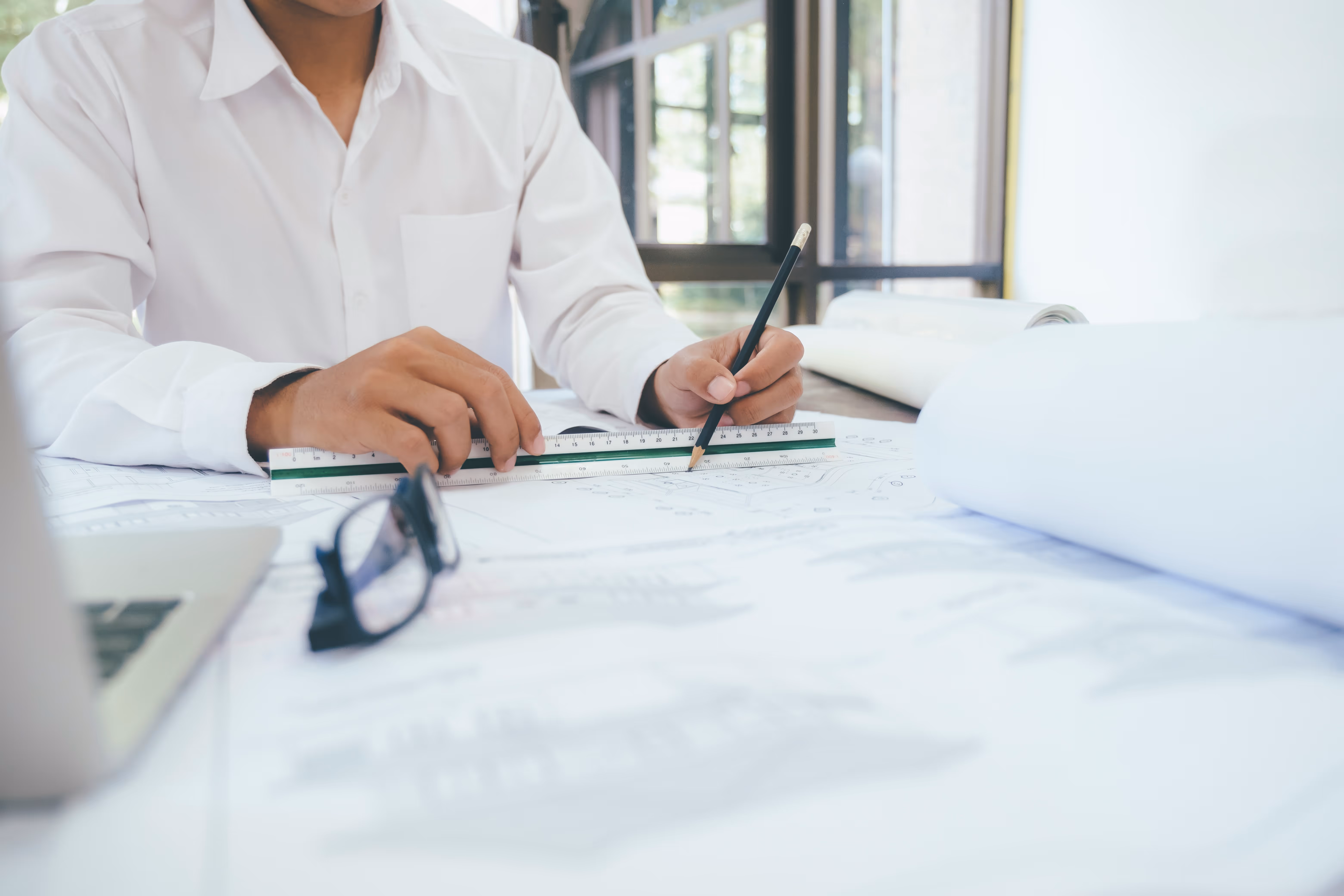 Person measuring architectural plans with a ruler and pencil on a table with blueprints and glasses.