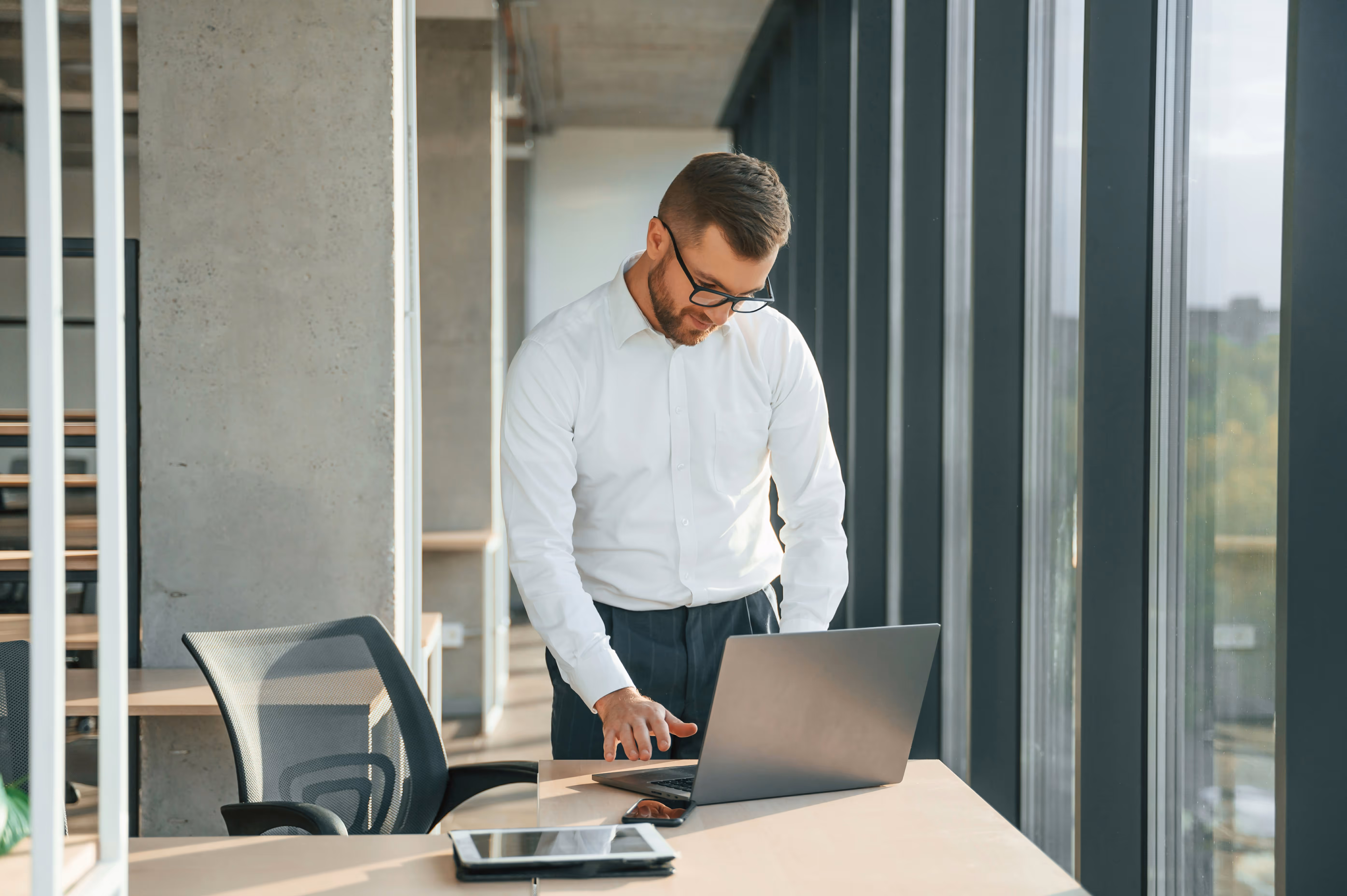 Homme en chemise blanche utilisant un ordinateur portable debout près d'une fenêtre de bureau lumineuse.