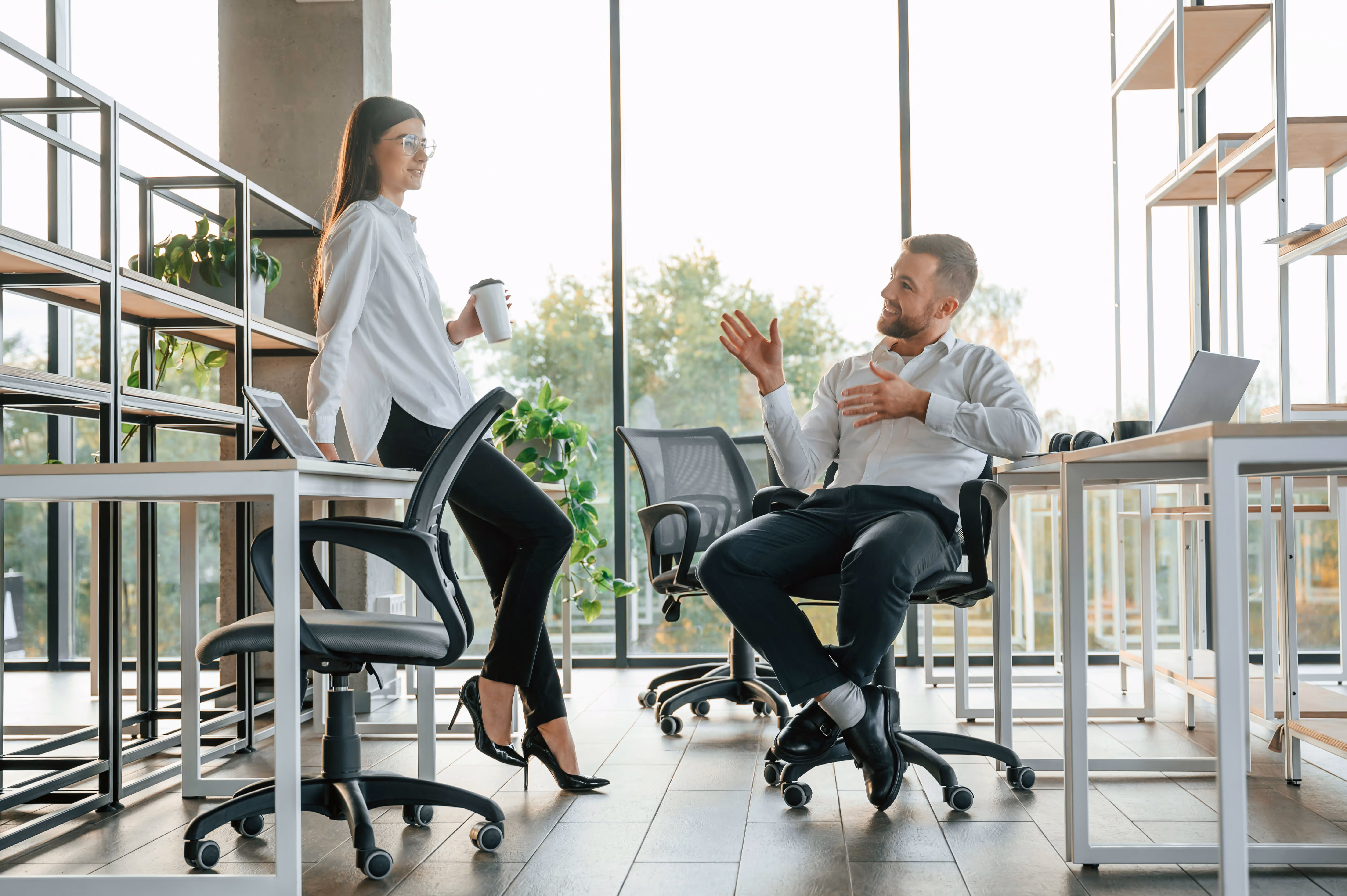 Deux collègues en blouse blanche discutent dans un bureau moderne avec grandes fenêtres et plantes.