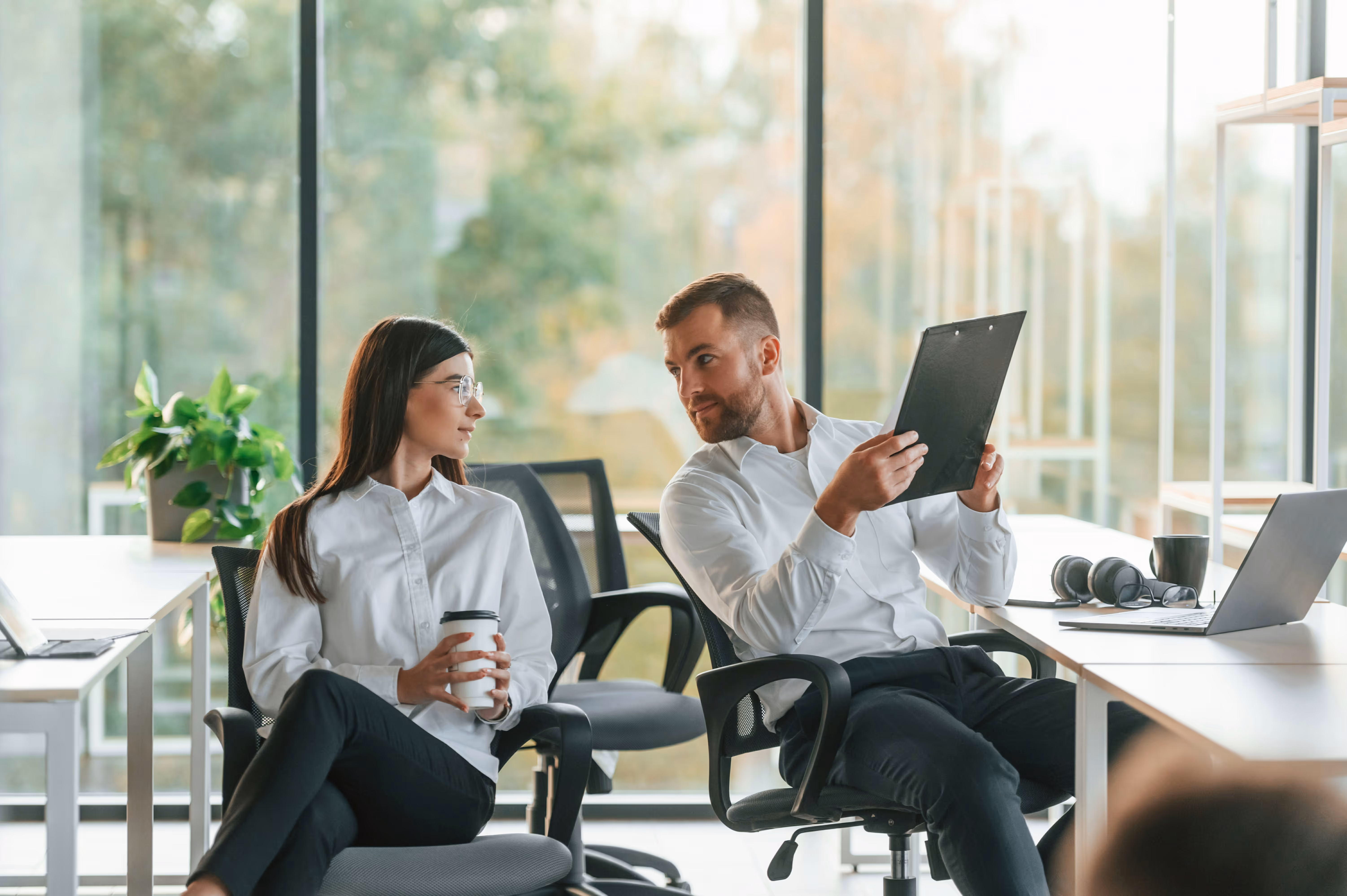 Deux collègues en chemises blanches discutent dans un bureau moderne avec fenêtres et laptop.
