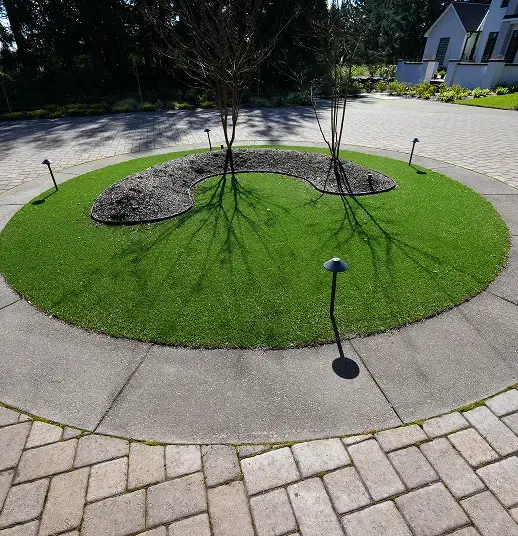 Circular patch of artificial grass with a kidney-shaped mulch bed and small trees in a paved driveway.