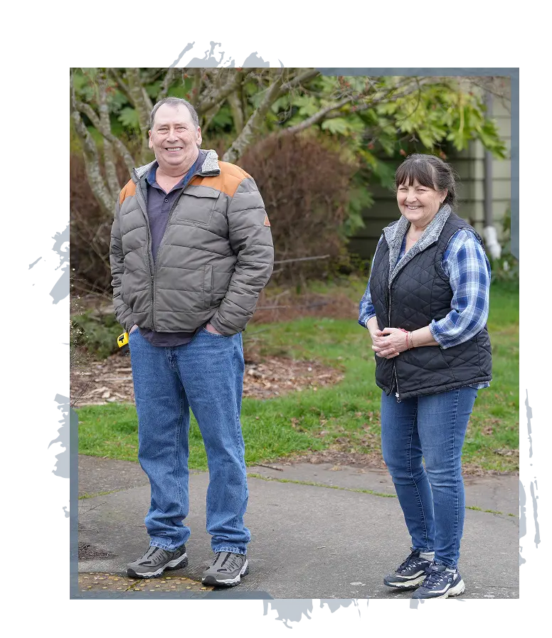 Smiling man and woman standing outdoors on a pathway with trees and grass in the background.