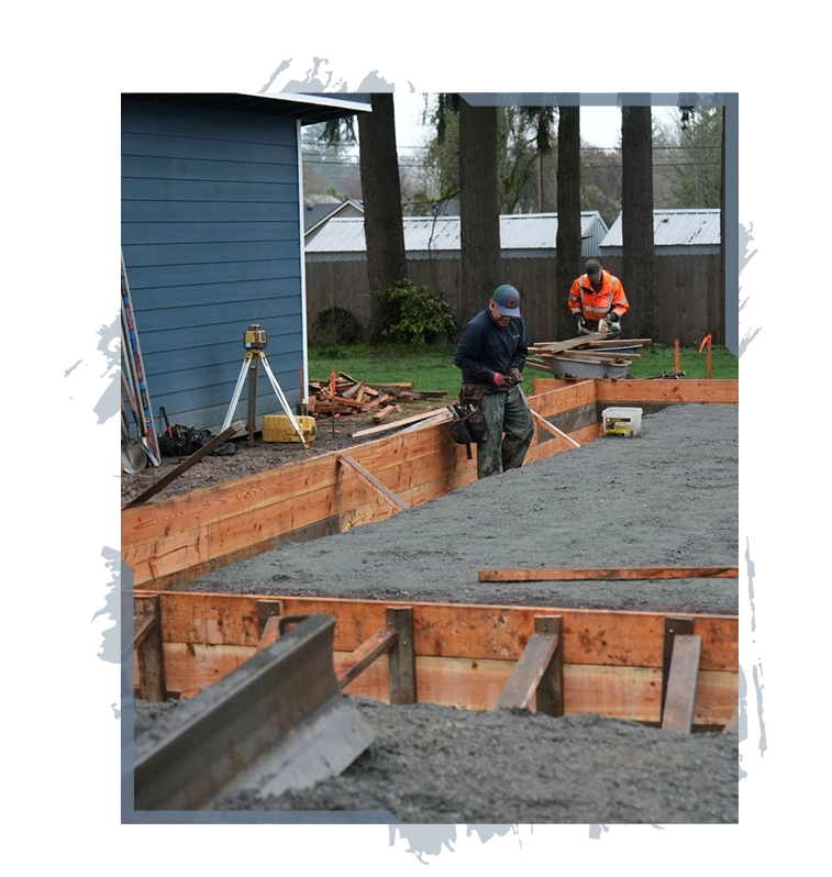Two construction workers preparing wooden forms and leveling poured concrete outside a blue building.