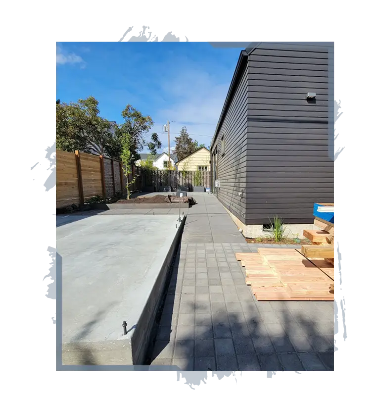 Concrete foundation slab next to a dark wooden house with paving stones and wooden planks nearby.