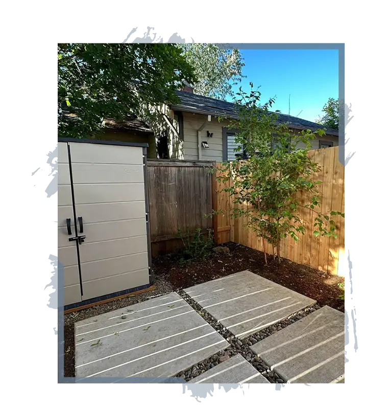 Small backyard with paver stone path, storage shed, young tree, and wooden fence under clear sky.