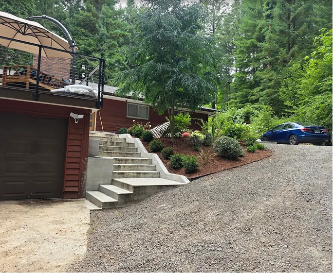 Gravel driveway leading to a red house with concrete stairs and garden beds with shrubs and trees.