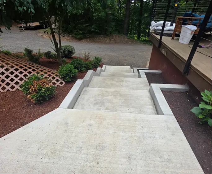 Concrete stairs leading down with landscaping and porch on either side in outdoor wooded area.
