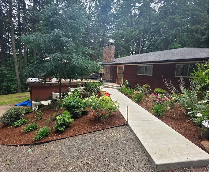 Concrete walkway leading to a brown house surrounded by flower beds and trees in a forested area.