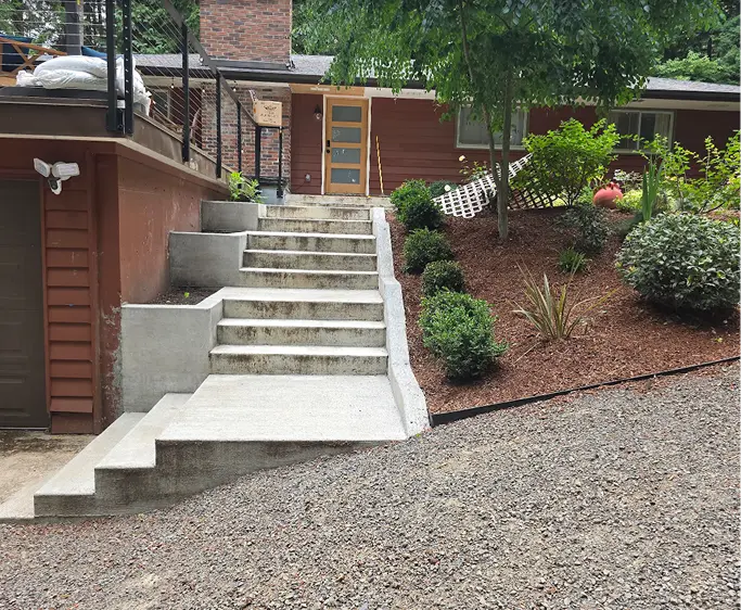 Concrete stairs with landings leading up to a front door beside a landscaped yard with bushes and trees.