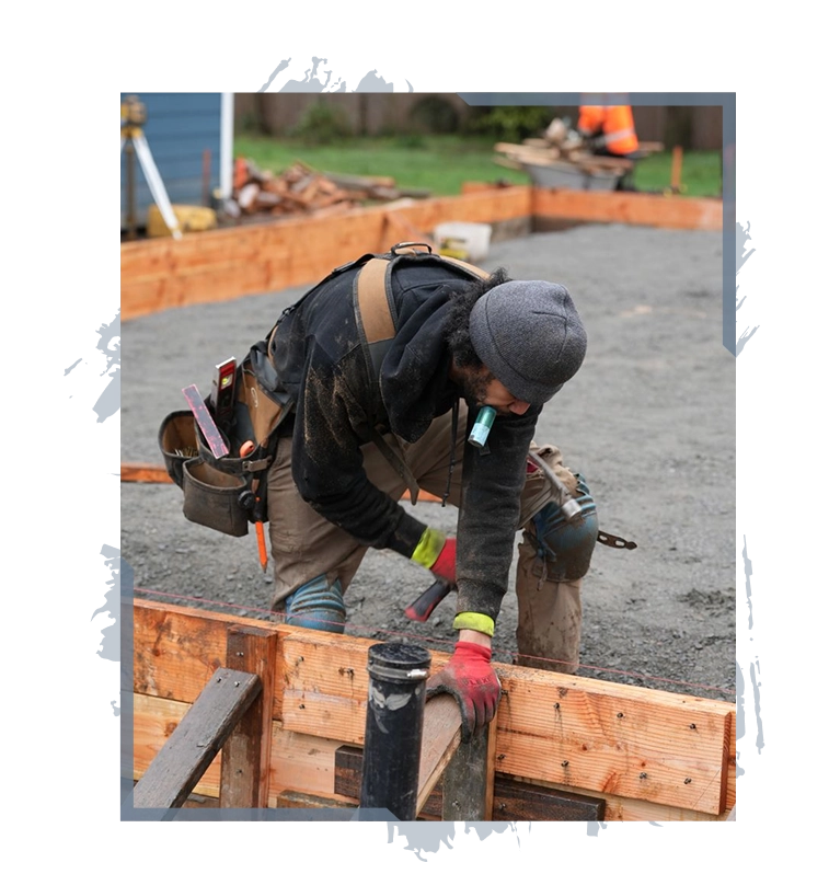 Construction worker wearing a beanie and gloves hammering wood frame on a gravel site.