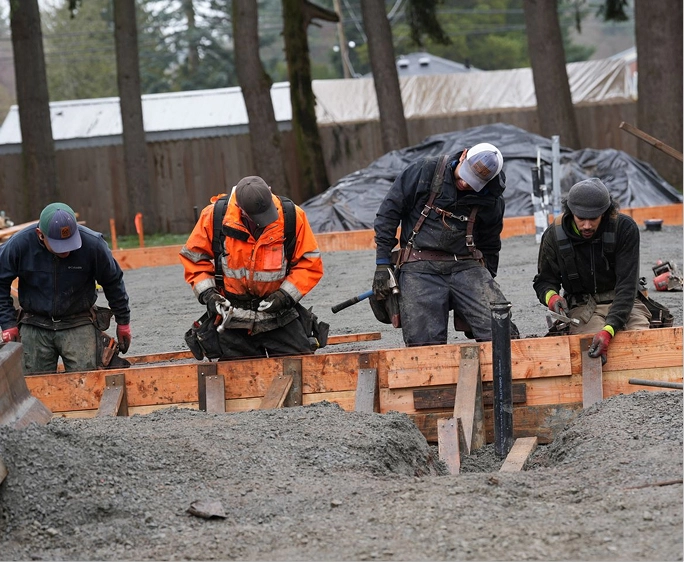 Four construction workers smoothing concrete within wooden framing outdoors near trees.