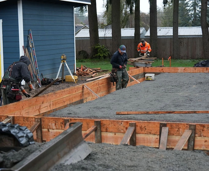 Workers preparing wooden forms and smoothing wet concrete for a building foundation next to a blue house.