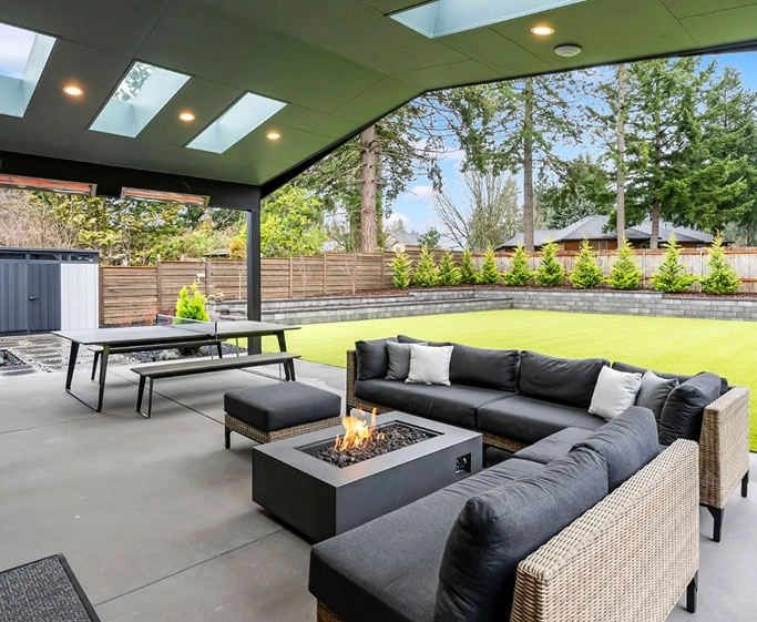 Covered patio with gray cushioned sectional, fire table, ping pong table, and green lawn beyond.