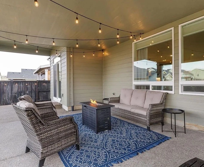 Covered patio with wicker sofa, chairs, blue rug, fire pit table, string lights, and side table.