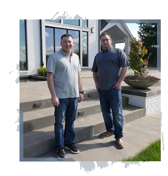 Two men in casual shirts and jeans standing on concrete steps outside a modern white brick house.