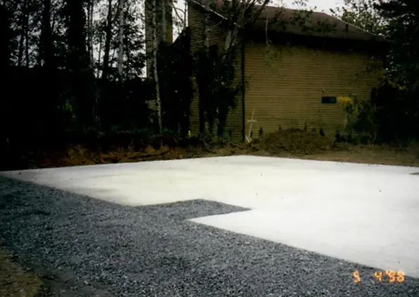 Concrete slab foundation bordered by gravel near a wooded area and a small wooden building.