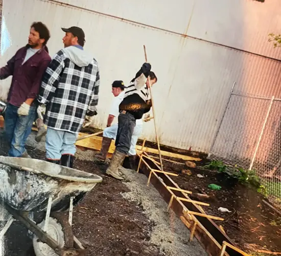 Four men working on concrete foundation next to metal fence and white wall with wheelbarrow nearby.