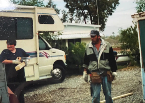 Two men outdoors near an RV and a small building, both wearing work gloves and casual clothes.