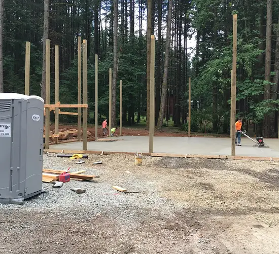 Construction site with wooden poles on concrete slab and workers preparing the ground in a forested area.