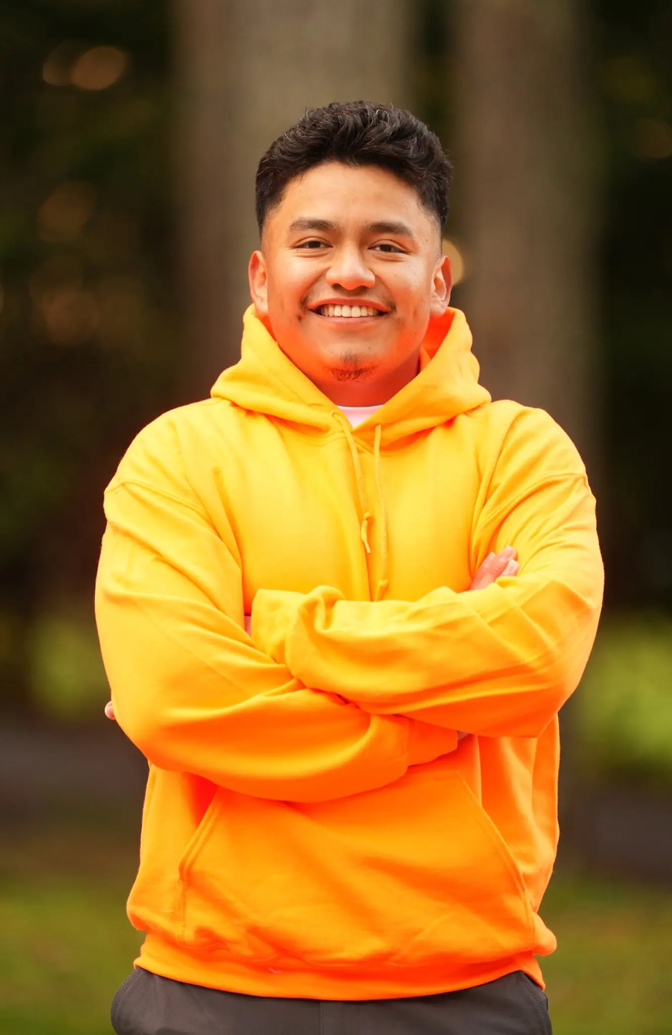 Smiling young man with short hair in a bright yellow hoodie standing outdoors with arms crossed.