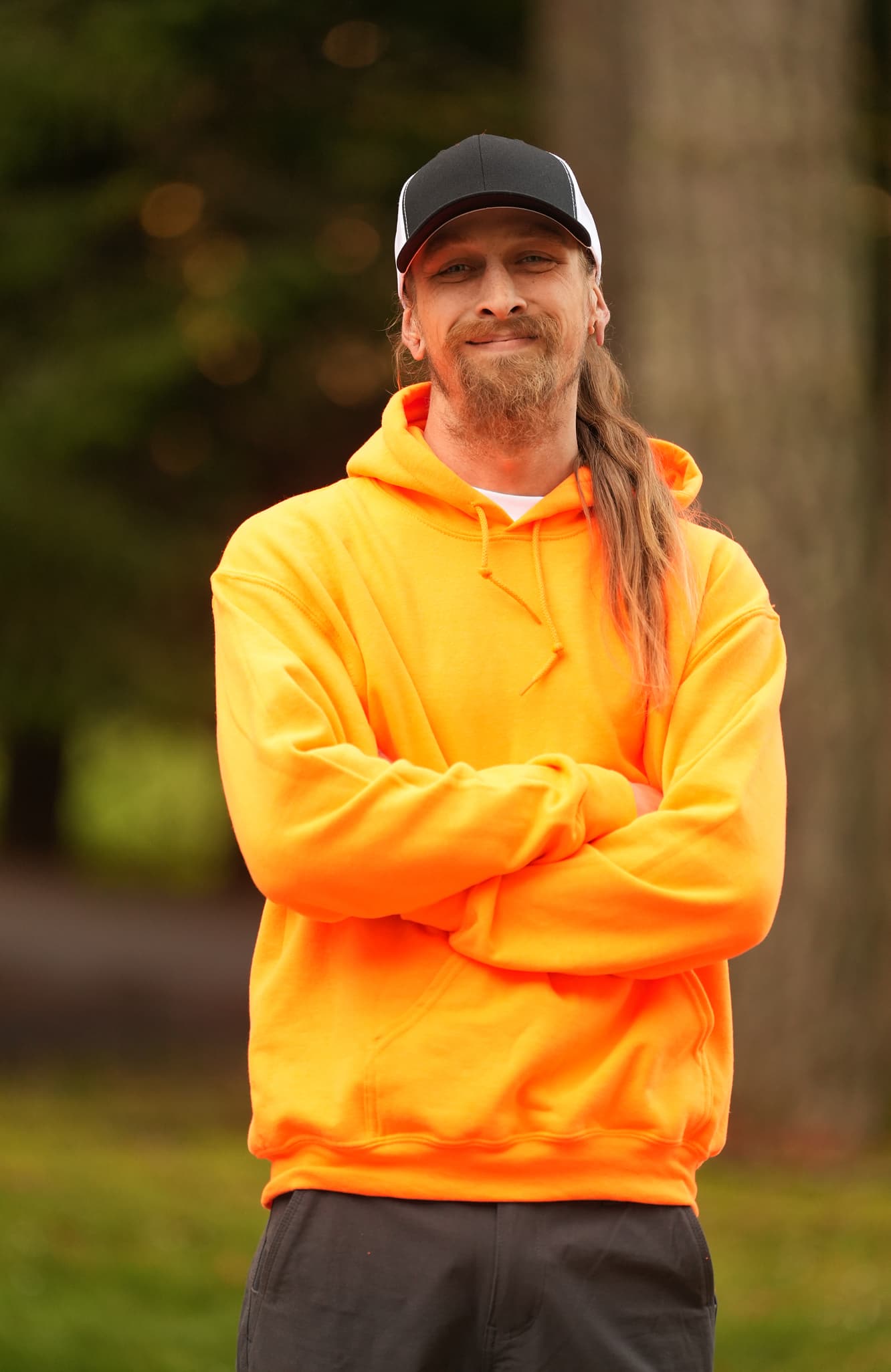 Man with long beard and hair in an orange hoodie and black cap stands outdoors with arms crossed.