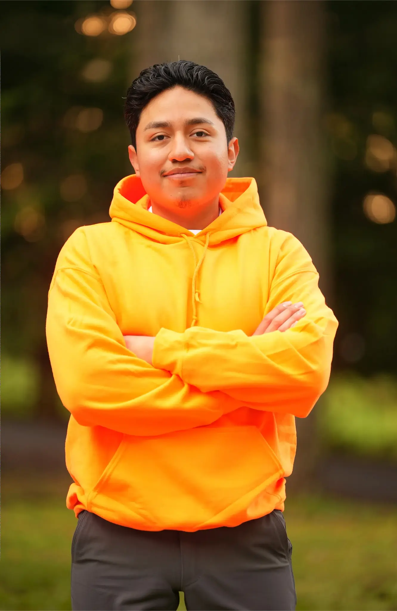 Man standing outdoors with arms crossed wearing bright orange hoodie and dark pants.