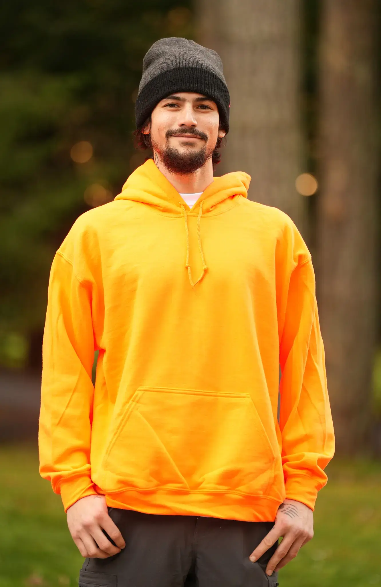 Man with beard and black beanie wearing bright orange hoodie standing outdoors with trees in background.