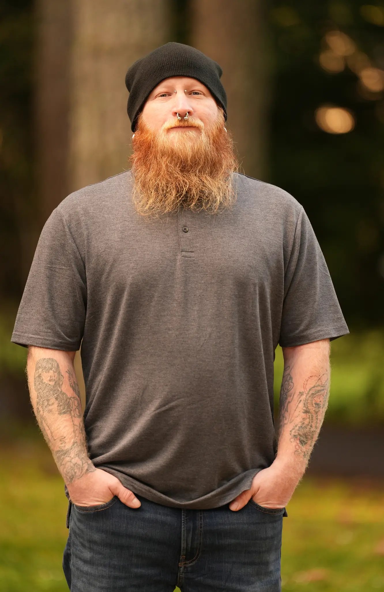 Bearded man in a black beanie and gray shirt with tattoos on his arms stands outdoors with hands in pockets.