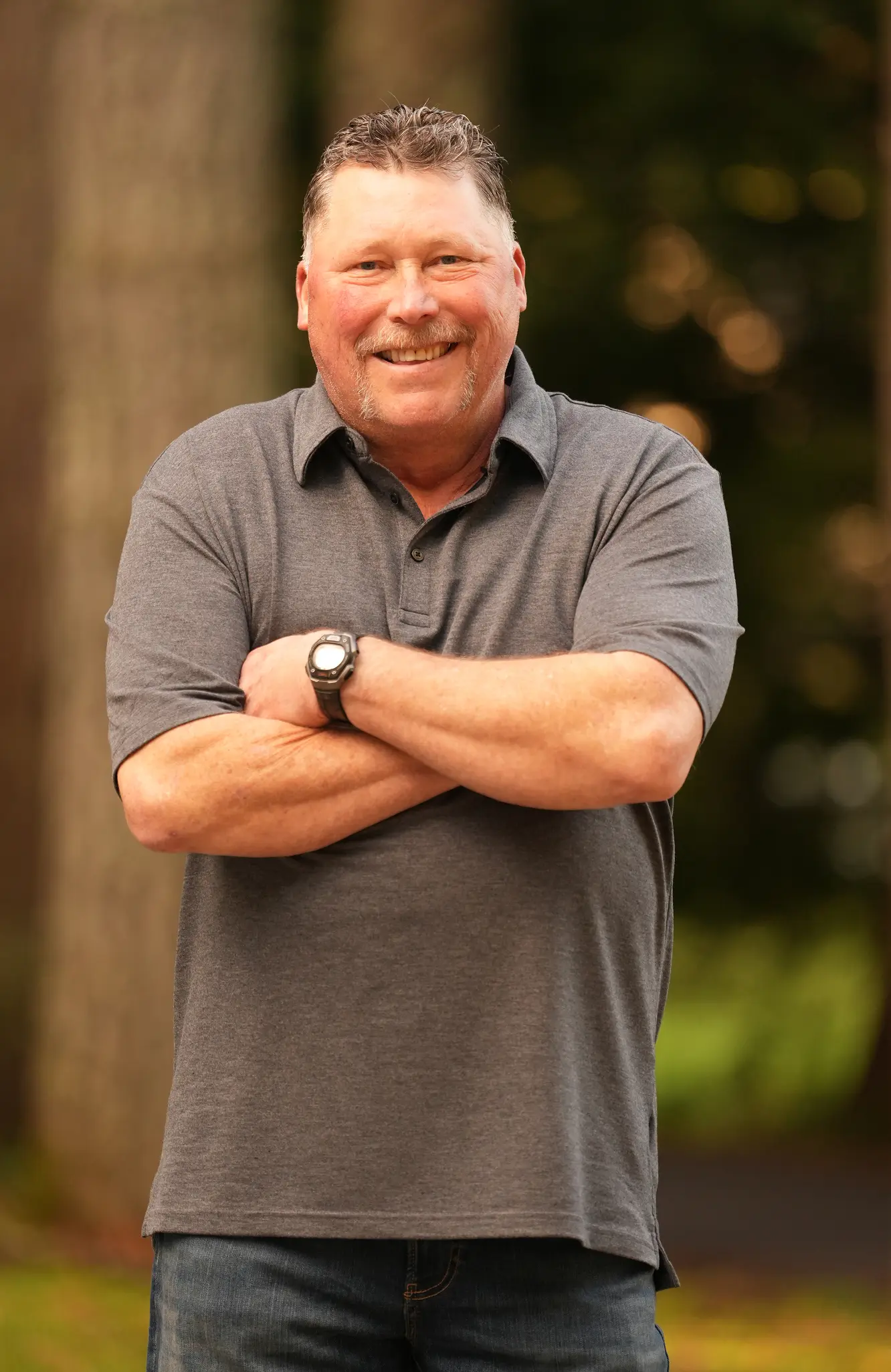 Smiling man with mustache in gray polo shirt and watch standing outdoors with arms crossed.
