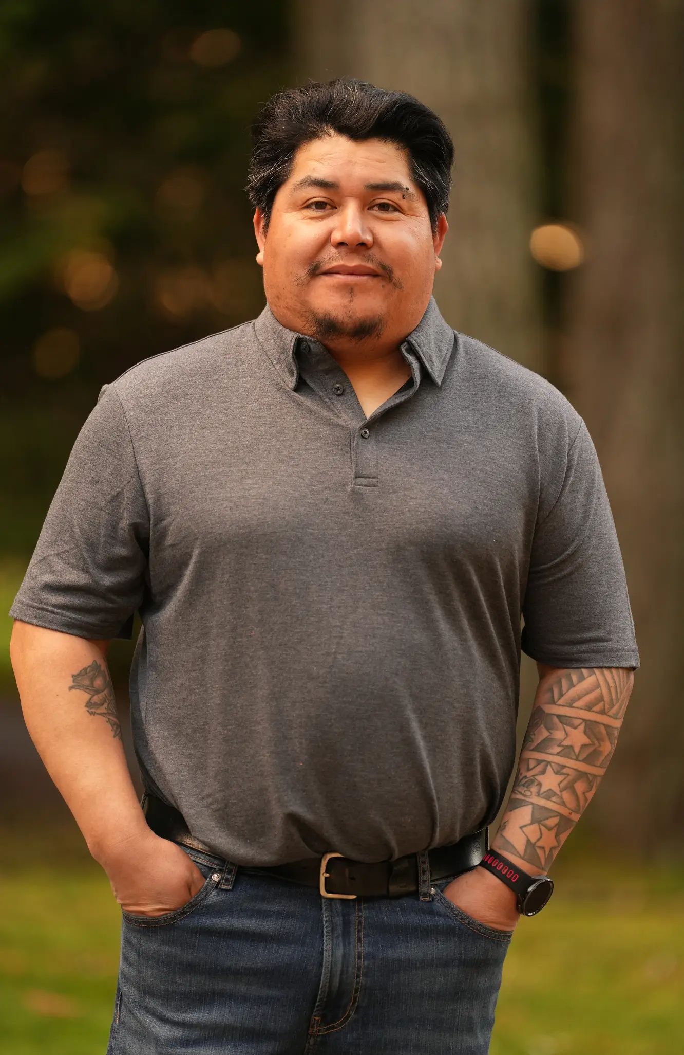 Man in gray polo shirt with tattoos on arms stands outdoors with hands in pockets.
