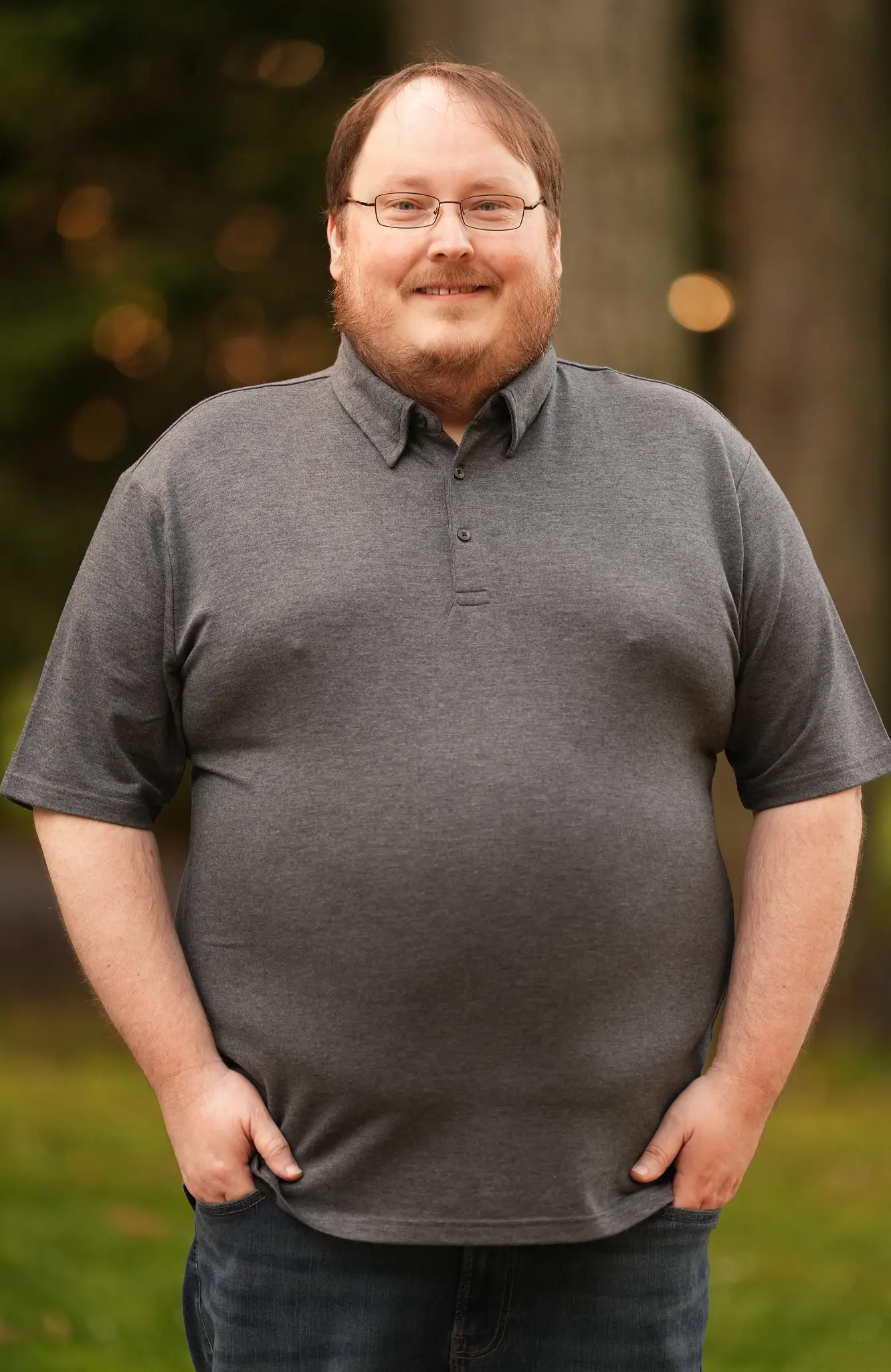 Smiling man with glasses and beard wearing a grey polo shirt and jeans standing outdoors.