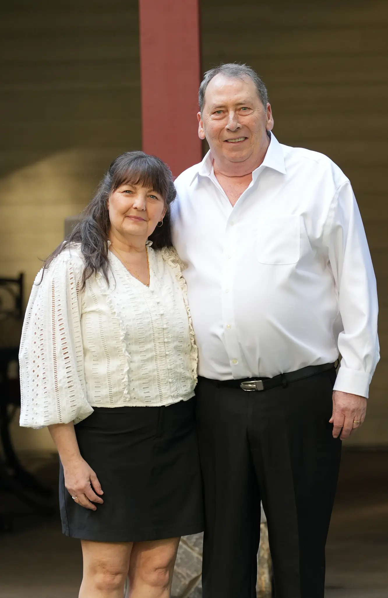 Smiling middle-aged couple standing together, man in white shirt and black pants, woman in white blouse and black skirt.