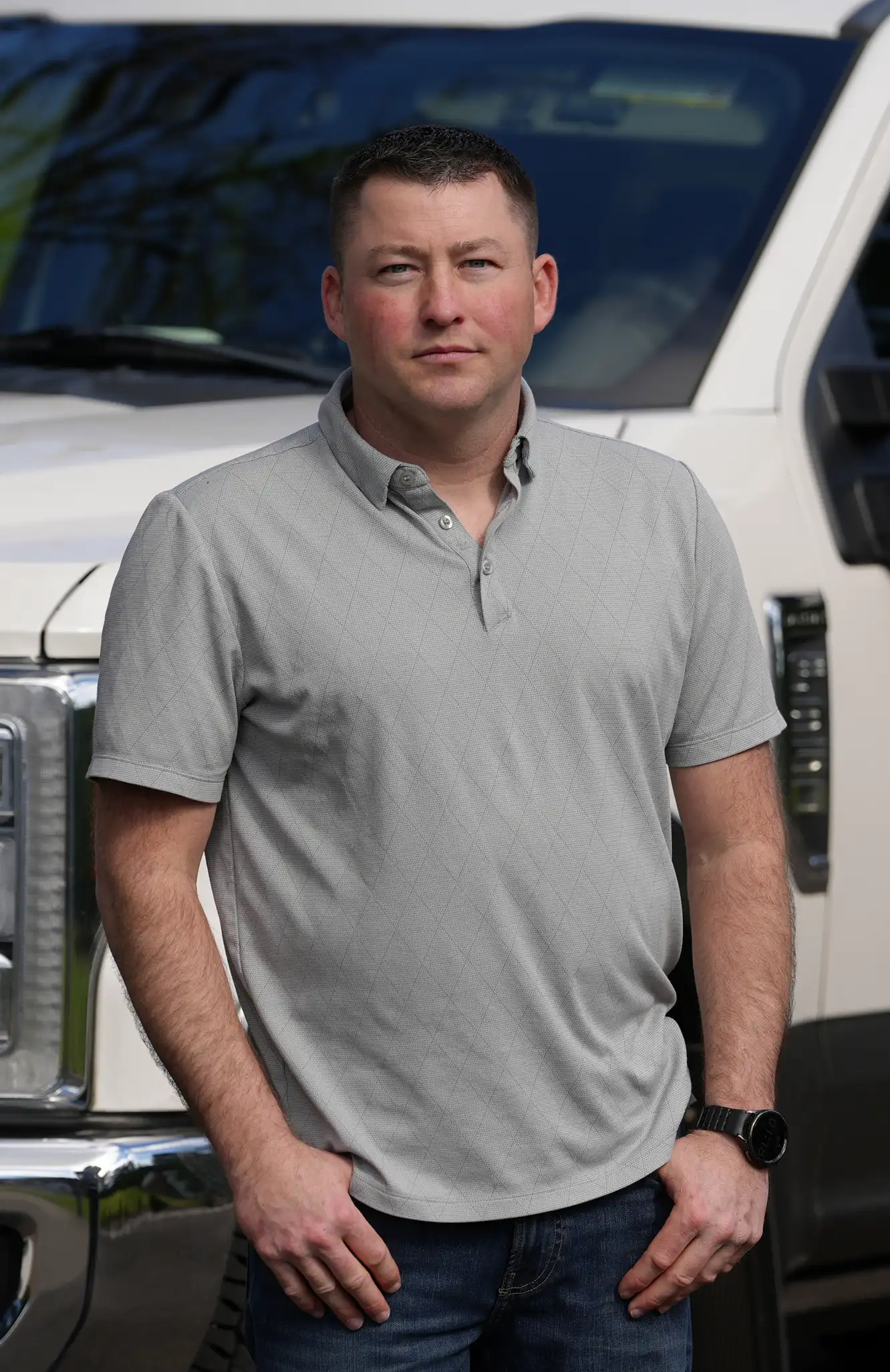 Man in gray polo shirt and jeans standing with hands in pockets in front of a white truck.