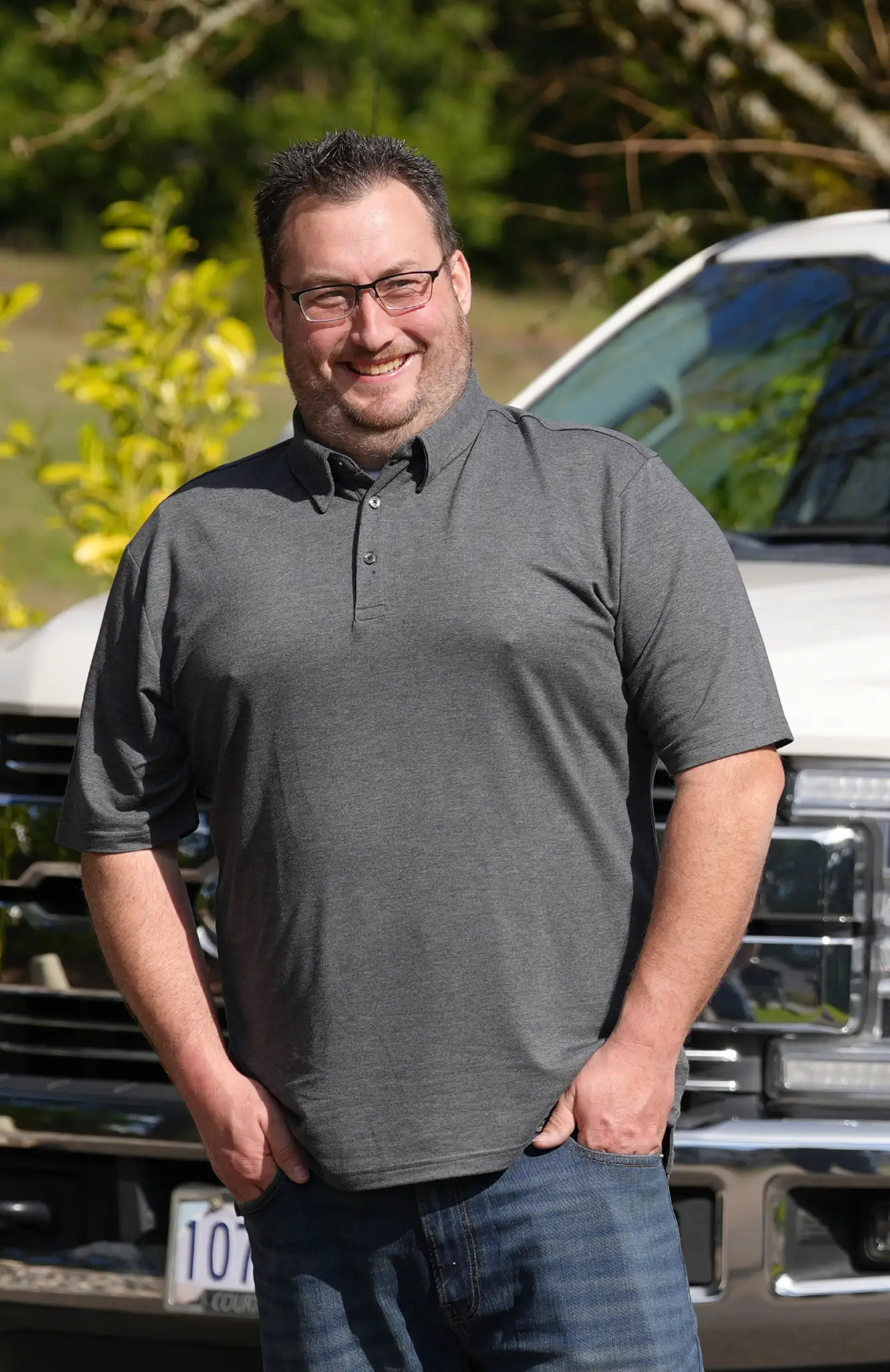 Smiling man in glasses and gray polo shirt stands with hands in pockets in front of a white truck.