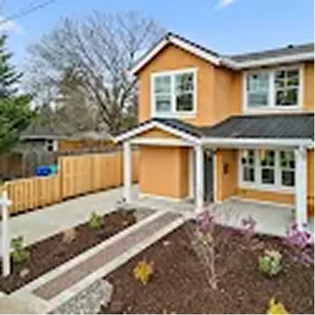 Two-story light orange house with a front porch, garden beds, and paved walkway under a blue sky.
