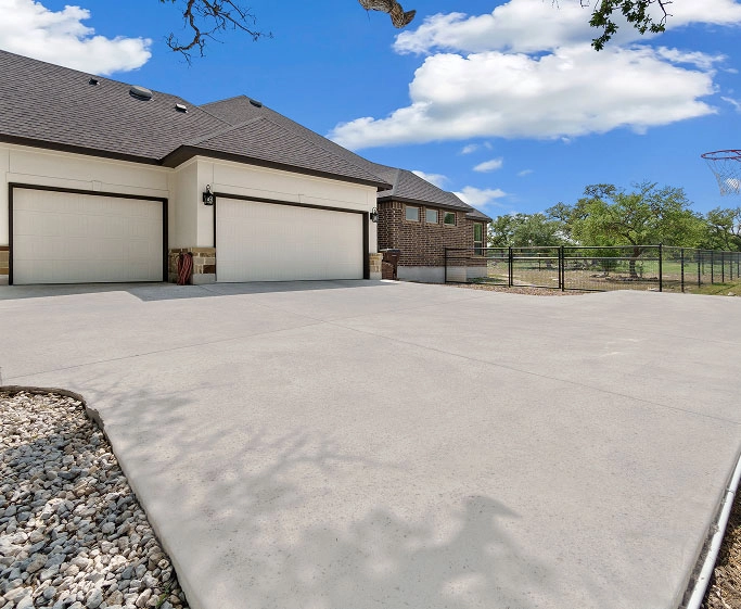 Wide concrete driveway leading to two closed garage doors on a house under a blue sky.