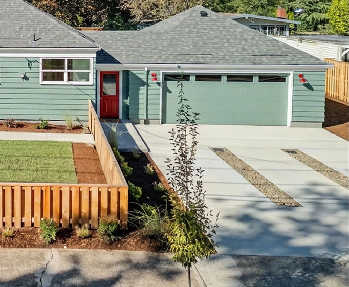 Green house with gray roof, red front door, fenced yard, and concrete driveway with gravel insets.