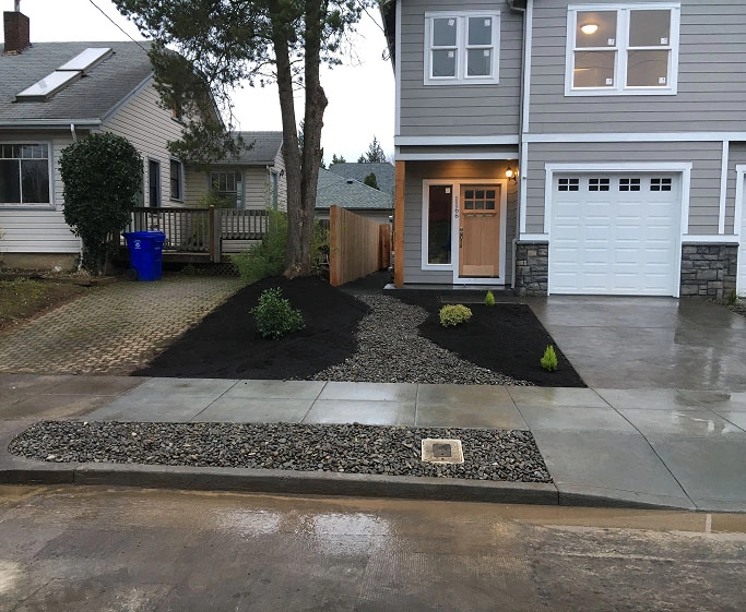 Modern gray house with wooden door, single garage, stone accents, and fresh landscaping with mulch and gravel path.