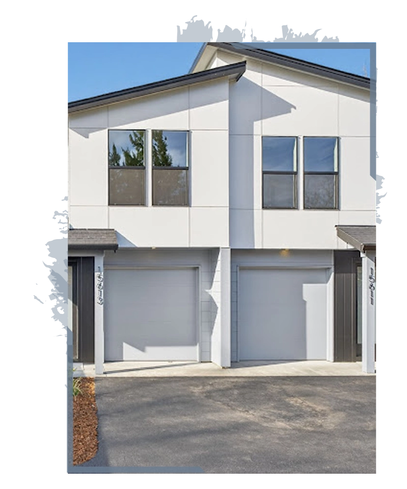 Modern duplex house with two garage doors, large windows, and a paved driveway under clear sky.