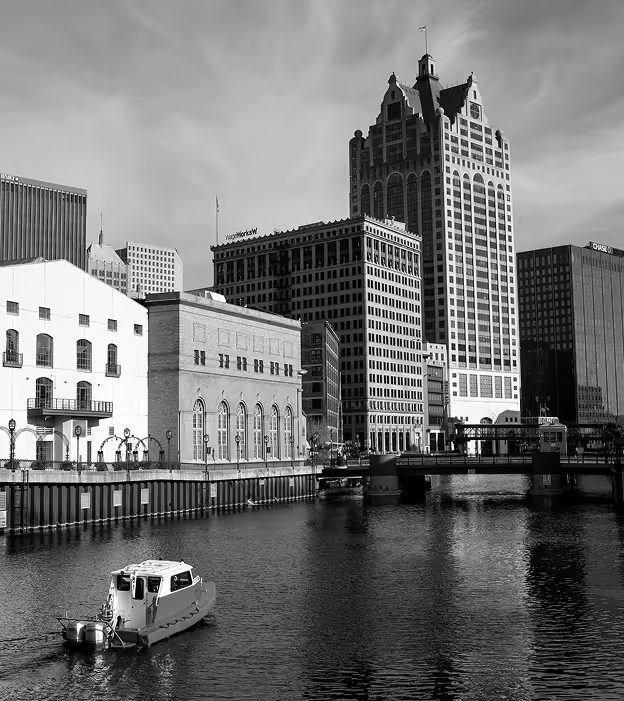 Black and white cityscape with river, small boat in water, bridge, and tall buildings in the background.