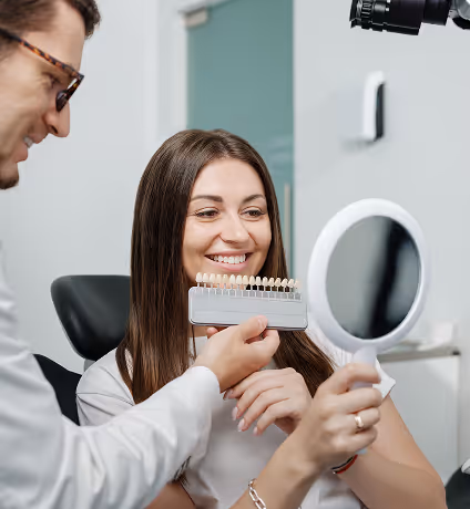 Dentist showing a smiling woman a dental shade guide while she holds a round mirror.