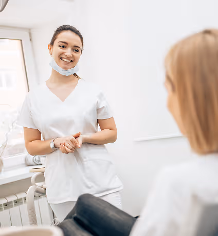 Smiling female healthcare professional with a mask under her chin talking to a seated patient in a bright room.