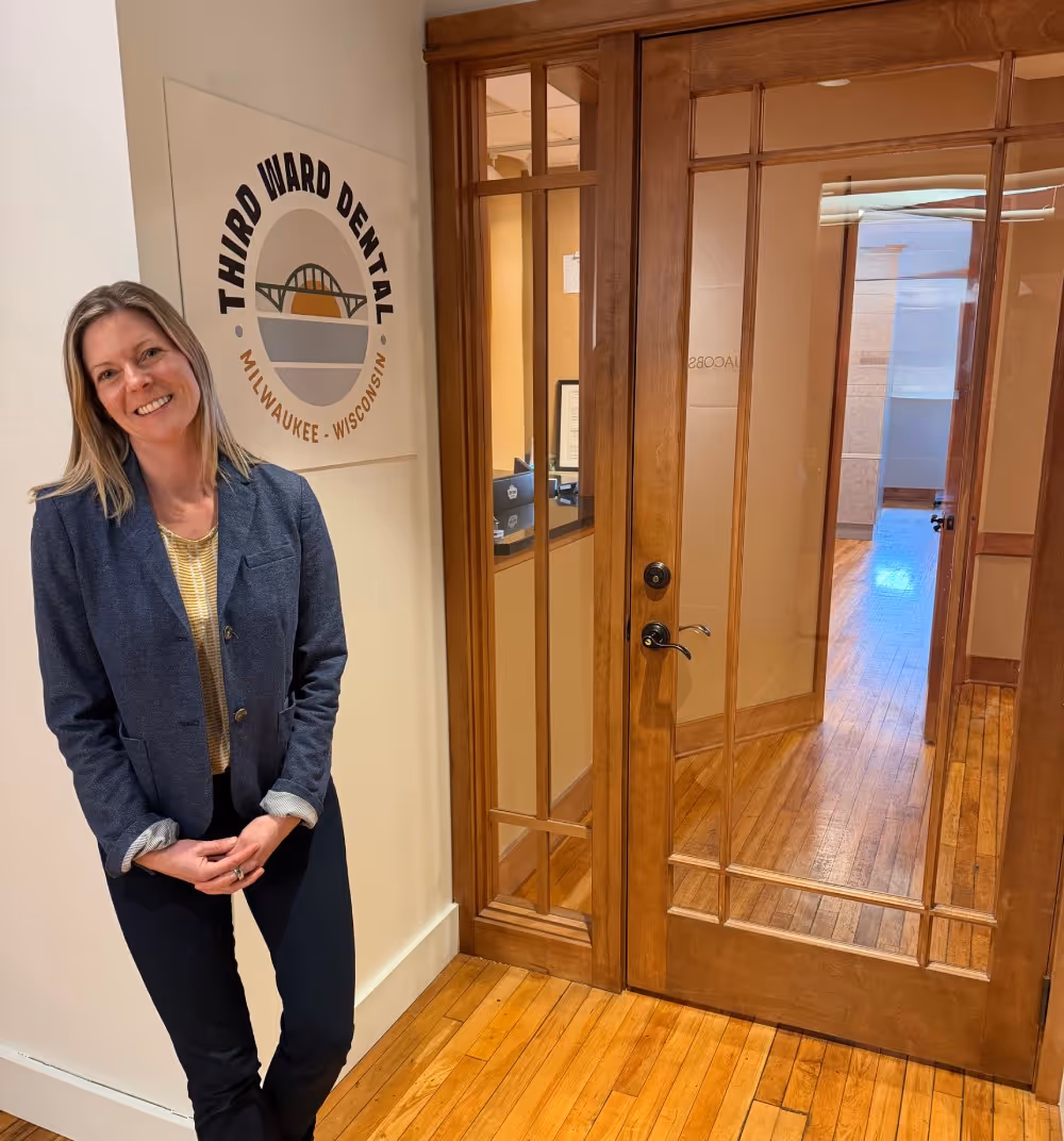 Smiling woman in blazer standing inside a dental office near a sign reading Third Ward Dental.