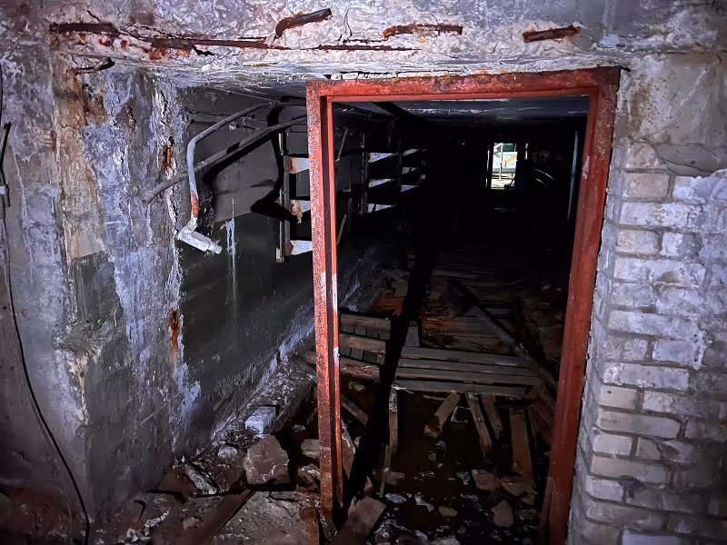 Dark, abandoned tunnel with broken wooden planks, rubble, and a red metal frame doorway.
