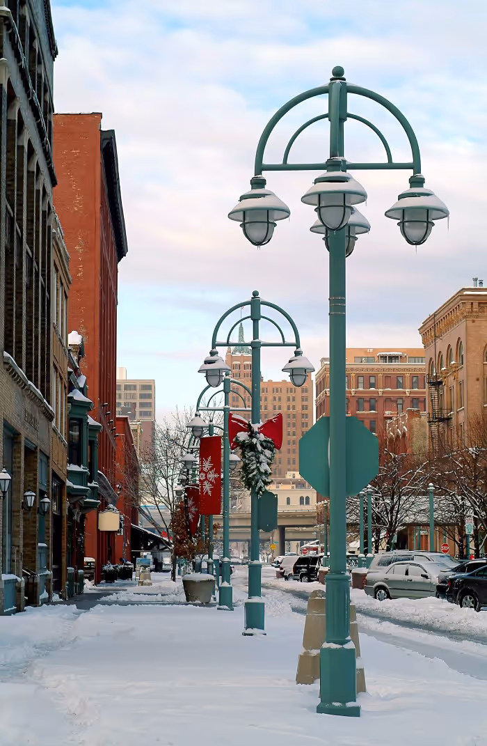Snow-covered city street with green lampposts decorated for Christmas and buildings in the background.