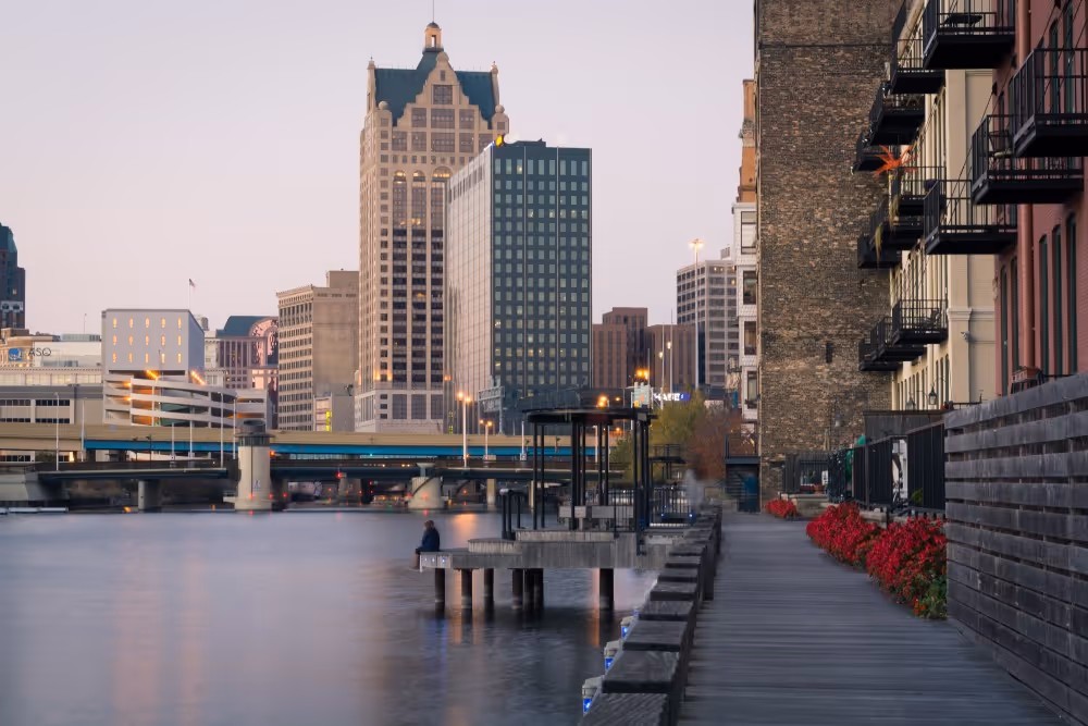 Riverwalk with red flowers and benches beside a city skyline during early evening.