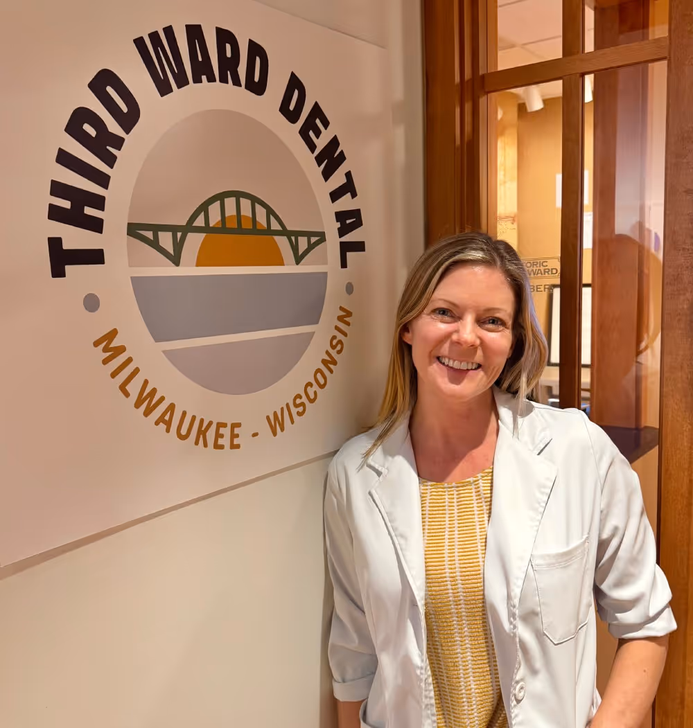 Smiling woman in a white coat stands next to a Third Ward Dental Milwaukee Wisconsin logo on a wall.