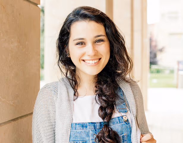 Smiling young woman with long braided hair wearing denim overalls and a light cardigan.