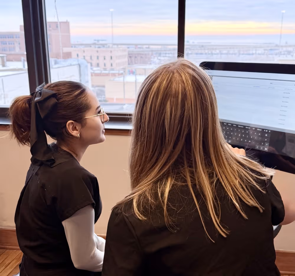 Two women in medical scrubs looking at a computer screen by a window with an urban view.