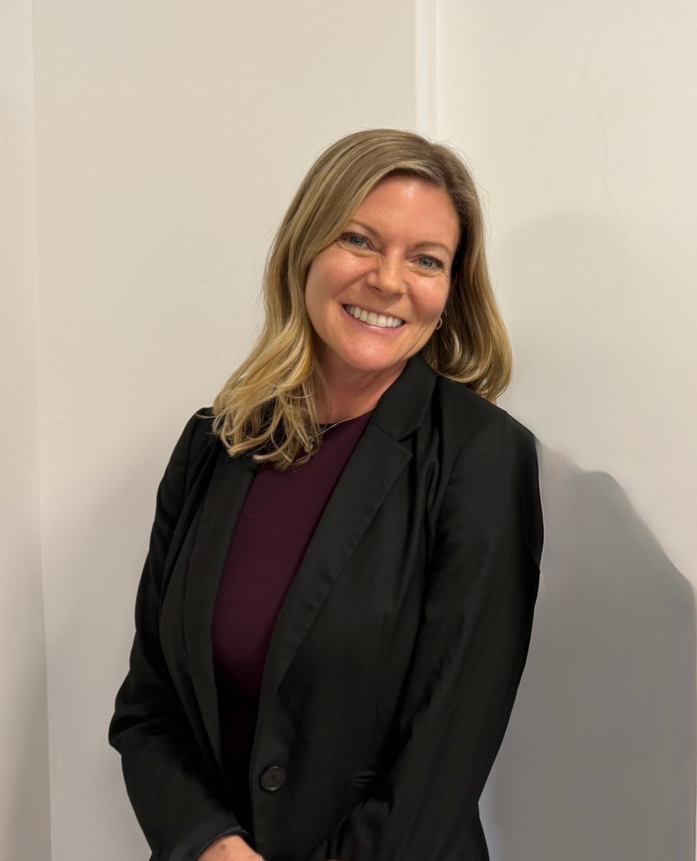 Smiling blonde woman wearing a black blazer and dark top standing against a white wall.